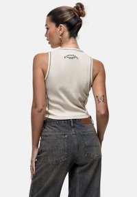 Top beige senza maniche con cuciture a contrasto e dettaglio del logo. Abbinato a jeans in denim scuro a vita alta con tasche posteriori e etichetta marrone.