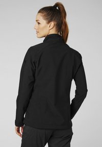 Chaqueta softshell negra con un alto cuello, que presenta una textura suave y una marca discreta en la manga. Diseño ajustado con costuras estructuradas.