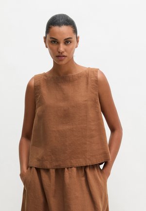 Femme les mains dans les poches, portant un haut sans manches en lin marron et un pantalon large assorti, sur un fond clair uni.