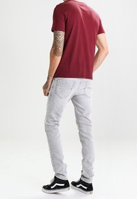 Burgundy t-shirt, korta ärmar, grå slim-fit jeans, detaljerad sömnad, svarta högsnabbasneakers med vita accenter, bäraren vänd bort.