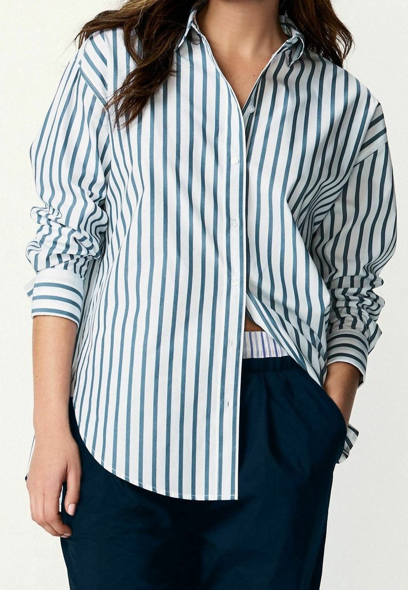 Femme portant une chemise blanche boutonnée avec des rayures verticales bleues, manches retroussées, et un pantalon bleu foncé avec une main dans la poche.