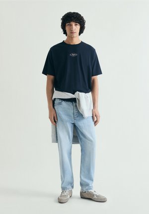 Joven con pelo rizado, camiseta negra, vaqueros azul claro, zapatillas grises y sudadera gris atada alrededor de la cintura.