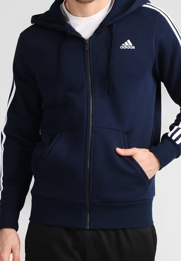Homme portant un sweat à capuche zippé bleu marine Adidas avec des bandes blanches sur les manches et un logo sur la poitrine, main dans la poche avant.
