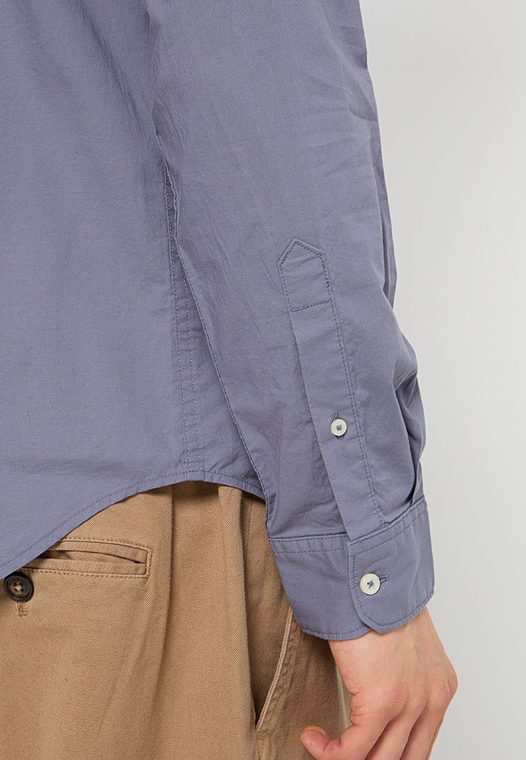 Camisa ajustada de color púrpura claro con cuello puntiagudo, mangas largas y puños asegurados con botones. Textura suave, dobladillo doblado y costuras bien hechas.