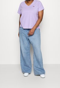 Liliowa bawełniana koszulka z dekoltem w serek z małym logo, połączona z jasnoniebieskimi szerokimi spodniami i białymi butami, stojąca na jednolitym tle.