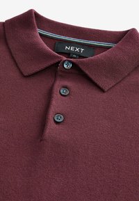 Polo en bourgogne avec une texture tricotée, trois boutons sombres et un col intérieur rayé. L'étiquette de marque "NEXT" est visible.