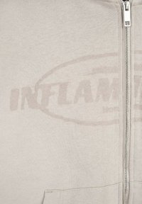 Jasnoszara bluza z kapturem na zamek, wykonana z miękkiego materiału. Posiada przednią kieszeń, metalowy zamek oraz delikatny nadruk "INFLAMMABLE" na klatce piersiowej.