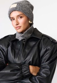 Căciulă gri tricotată cu etichetă, pulover gri cu guler înalt asortat și geacă de piele neagră cu detalii pe manșete ripate. Model cu brațele încrucișate.