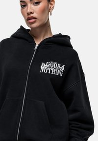 Felpa nera con zip in tessuto morbido, caratterizzata da un design grafico frontale con testo "GOOD NOTHIN'" e dettagli a forma di farfalla, grande cappuccio e tasche frontali.