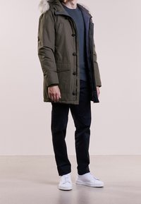 Oliwkowa parka z kapturem podszytym futrem, zapinana na guziki z dwoma bocznymi kieszeniami, w zestawie z czarnymi spodniami i białymi trampkami.