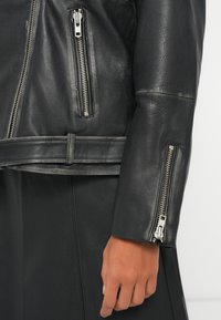 Μαύρο δερμάτινο τζάκετ με πολλές ασημένιες φερμουάρ, υφή και εφαρμοστή σιλουέτα με λεπτομέρεια στη μέση που ζώνεται.