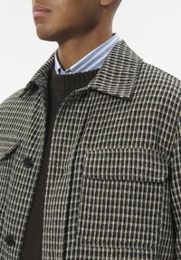 Ung mand iført en sort og beige ternet jakke over en mørkebrun sweater og en blåstribet skjorte med krave.