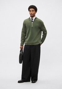 Πλεκτό πουλόβερ σε ελιά πράσινο με φερμουάρ στον γιακά, συνδυασμένο με μαύρο παντελόνι φαρδιάς γραμμής. Το μοντέλο κρατά μια μαύρη δερμάτινη τσάντα και φορά μαύρα παπούτσια.