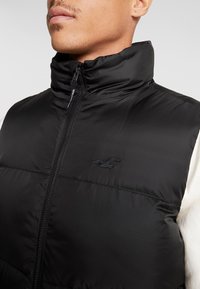 Man som bär en svart puffer-väst med hög krage och en synlig sydd logotyp på vänster bröst över en vit långärmad skjorta.