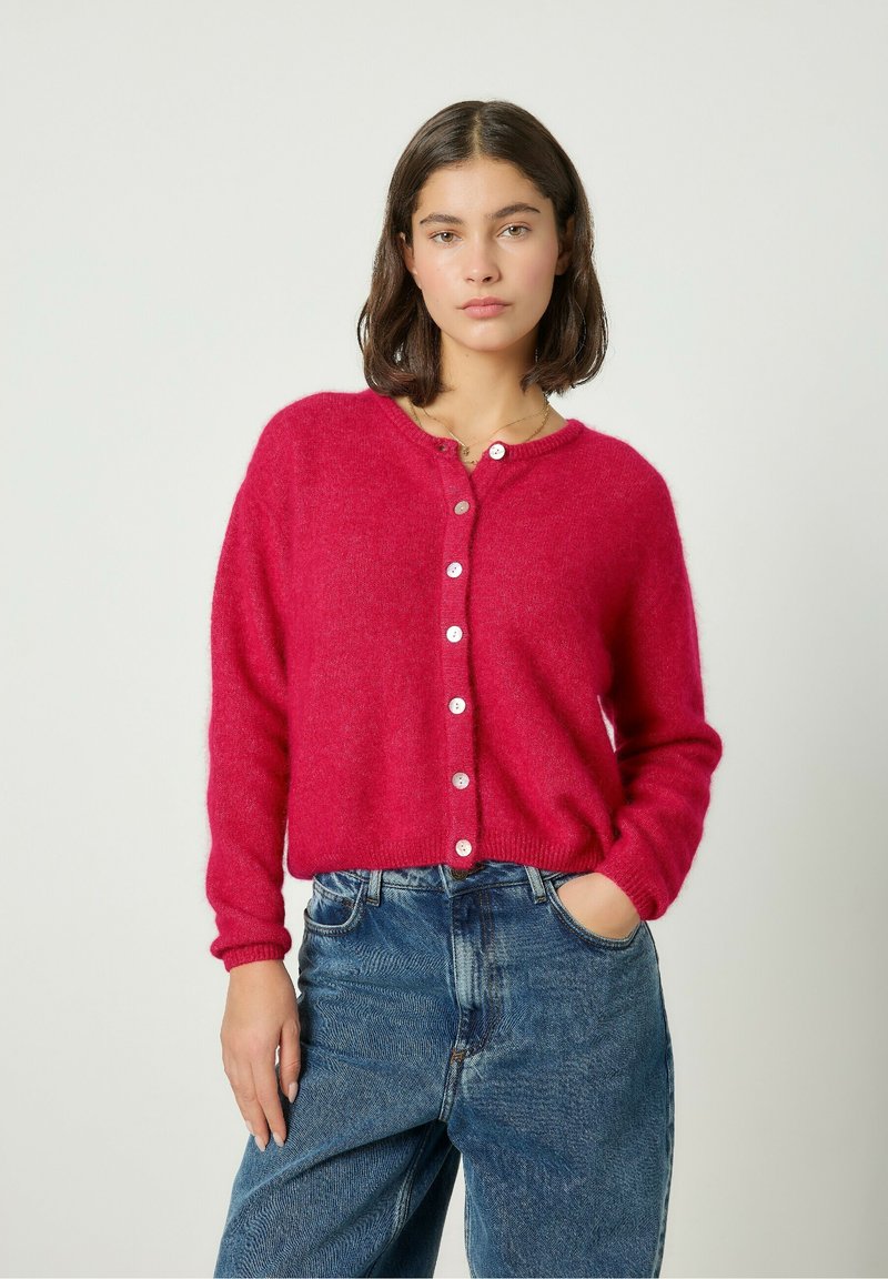 Cárdigan de punto fucsia con cuello redondo, botones frontales y puños acanalados, combinado con jeans de mezclilla de cintura alta.