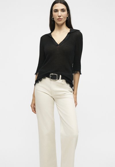 Γυναίκα που φοράει μαύρο τοπ με V-λαιμόκοψη και ξεφτισμένα άκρα και κρεμ παντελόνι με φαρδιά μπατζάκια, στέκεται μπροστά σε ουδέτερο φόντο.