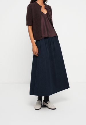 Kvinde iført en mørkebrun kortærmet cardigan over en matchende top, et langt navy nederdel, sorte sokker og lyse loafers.