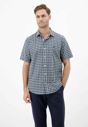 Mann trägt ein marineblau-weiß kariertes kurzärmeliges Hemd mit Knöpfen und dunkelblaue Hose, steht mit einer Hand in der Tasche.