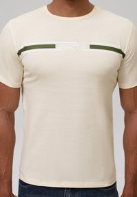 Camiseta de manga corta en color beige con cuello redondo. Presenta una franja horizontal verde y blanca con el texto "CRAFTED GOODS EST. 1958."