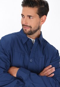 Camicia blu navy con colletto strutturato, pieghe frontali e cuciture a contrasto. Presenta due bottoni sui polsini e una texture liscia.