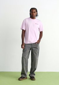 Pink short-sleeve t-shirt with "Dickies" logo, paired with loose-fitting gray work pants and dark shoes, standing on green flooring.