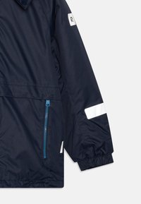 Giacca blu navy con tasca con zip, polsini elasticizzati, dettagli riflettenti bianchi e una patch con il logo sulla manica. Tessuto liscio e resistente alle intemperie.