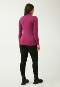 Figurbetonter magentafarbener Rollkragenpullover aus weichem Material, mit langen Ärmeln, einem gerippten Saum und einer glatten Textur. Getragen mit schwarzen Leggings und Stiefeln.