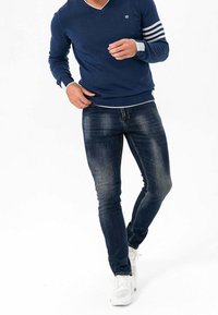 Marineblauer Pullover mit weißen Streifen an den Ärmeln, kombiniert mit dunkelblauen Jeans. Das Model trägt weiße Sneakers und präsentiert eine legere, figurbetonte Silhouette.