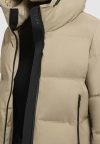Beżowa kurtka puchowa z wysokim kołnierzem, czarnym zamkiem błyskawicznym i bocznymi kieszeniami. Posiada pikowane wzory i gładkie, matowe wykończenie.