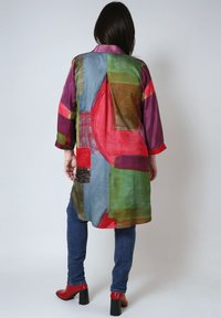 Frau in einem bunten, abstrakt gemusterten langen Mantel, blauen Jeans und roten, knöchelhohen Schuhen mit Absatz, die vor einem schlichten, hellen Hintergrund steht.