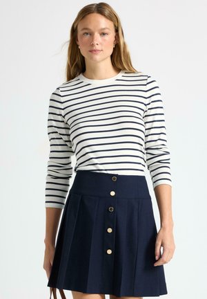 Top a maniche lunghe color crema con strisce blu abbinate a una gonna plissettata blu con bottoni dorati davanti. Tessuto morbido con colori a contrasto.
