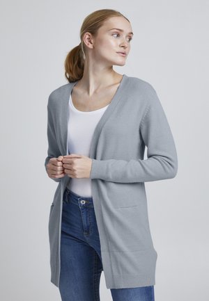 Ljust grå öppen kort cardigan, stickad textil, långa ärmar, framfickor, mjuk textur, som bärs tillsammans med en vit skjorta och blå jeans.