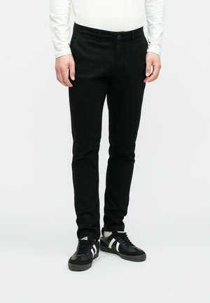 Man draagt strakke zwarte broek, zwart-witte sneakers en een wit shirt met lange mouwen, staand tegen een effen achtergrond.