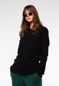 Schwarzes Sweatshirt mit langen Ärmeln und rundem Ausschnitt, kombiniert mit grünen Jogginghosen. Das Modell trägt dunkle Sonnenbrillen; die glatte Stoffstruktur ist sichtbar.