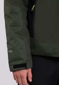 Giacca impermeabile verde caratterizzata da pannelli testurizzati e lisci, polsini regolabili con velcro e una tasca con zip dotata di un tirante giallo.