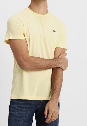 Homme portant un t-shirt jaune pâle à manches courtes et un jean bleu foncé, tenant son bras gauche avec sa main droite sur un fond blanc.