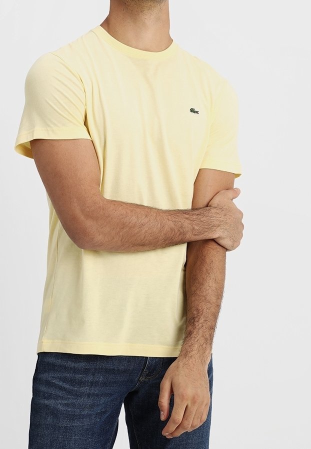 Homme portant un t-shirt jaune pâle à manches courtes et un jean bleu foncé, tenant son bras gauche avec sa main droite sur un fond blanc.
