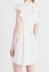 Nainen, jolla on yllään lyhyt valkoinen mekko, jossa on liehuvat hihat, teksturoitu kangas ja takana nappikiinnitys avokuja, kuvattu takaa.