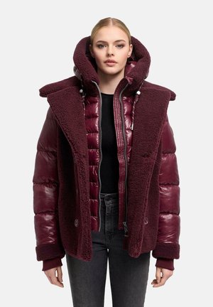 Jeune femme portant une veste matelassée bordeaux à plusieurs couches avec des panneaux en polaire sur un haut noir et un jean foncé sur un fond uni.