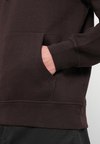 Ruka vo vrecku tmavohnedej mikiny, ktorá ukazuje detailné šitie a rebrovaný lem rukáva.