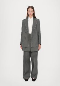 Grå overdimensjonert blazer med hvit søm, dobbeltknappet design, kombinert med grå vide bukser og en hvit skjorte med krage under.
