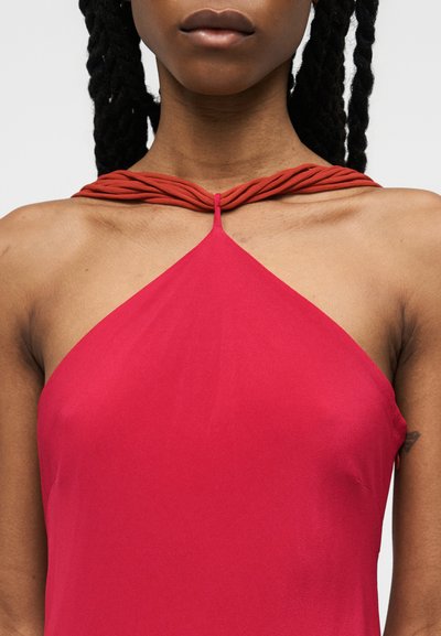 Νεαρή γυναίκα με σκούρα κοτσίδες που φοράει κόκκινο φόρεμα με λαιμόκοψη halter και στριφογυρισμένους ιμάντες γύρω από τον λαιμό, αφήνοντας ακάλυπτους τους ώμους και το άνω μέρος του στήθους.