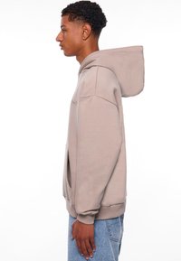 Lichtgrijze hoodie met een voorzak, gestructureerde mouwen en geribbelde manchetten, getoond op een model vanuit een zijaanzicht.