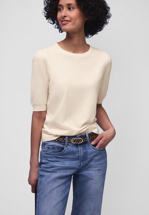 Kobieta o kręconych włosach, ubrana w kremowy sweter z krótkim rękawem, niebieskie dżinsy i pasek w panterkę, stojąca z jedną ręką w kieszeni.