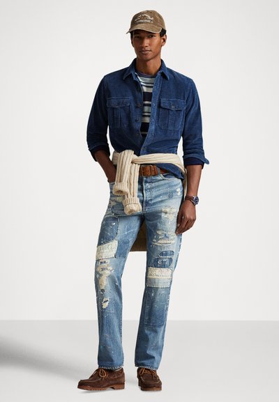 Vīrietis, kurš valkā zilu kordurojas kreklu virs svītraina T-krekla, nodilušas džinsu bikses ar pleķīšiem, bēša krāsas džemperi, sasietu ap vidukli, brūnas kurpes un brūnu cepuri.