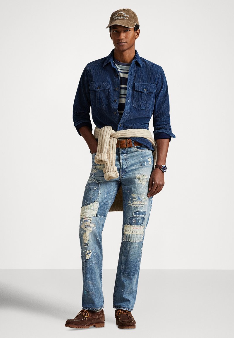 Uomo con una camicia di velluto a coste blu sopra una t-shirt a righe, jeans strappati con toppe, maglione beige legato in vita, scarpe marroni e cappello marrone.