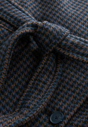 Kaput u vuni mješavini tamnoplave i smeđe boje s uzorkom houndstooth, s vezanom strukom i detaljem crnog dugmeta. Mekani, teksturirani materijal.