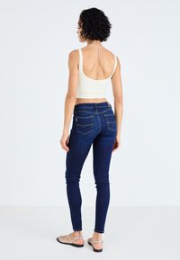 Fehér bordázott crop top, hátán kivágással, sötétkék skinny farmernadrággal párosítva, amely sárga varrással és ezüst lánc díszítéssel rendelkezik a nyakrészen.