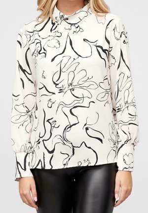 Blouse blanche à manches longues avec des lignes florales abstraites noires, portée avec un pantalon en cuir noir par une personne debout.