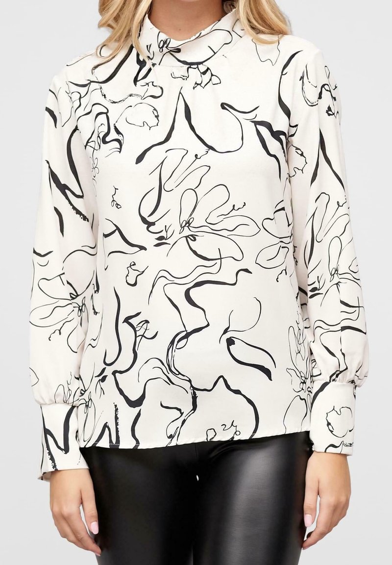 Blouse blanche à manches longues avec des lignes florales abstraites noires, portée avec un pantalon en cuir noir par une personne debout.
